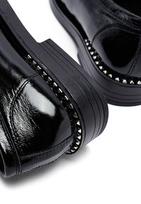 Zapatos de charol negros con suelas gruesas y acanaladas, adornados con tachuelas en forma de pirámide plateadas que bordean los extremos cerca del talón y la punta.