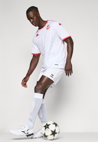 Balta futbola krekla ar sarkaniem akcentiem un logo, kopā ar baltām biksēm un zeķēm. Blakus ir melna un balta futbola bumba ar zvaigžņu rakstu.