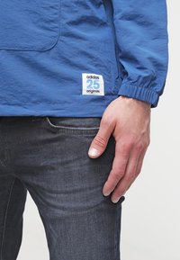 Modrá lehká bunda s přední kapsou, elastickými manžetami a značkovou nášivkou s nápisem "adidas 25 originals" na dolním lemu.