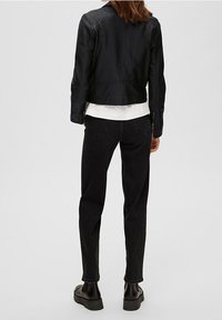 Schwarze Lederjacke mit figurbetontem Design, Reißverschlüssen an den Bündchen, über einem weißen Hemd getragen, kombiniert mit dunkler Jeans und klobigen schwarzen Stiefeln.