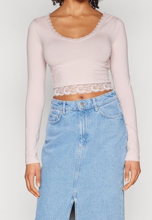 Kobieta ubrana w jasnoróżowy, długi crop top z koronkową lamówką i jasnoniebieską, wysoką spódnicę dżinsową z rozcięciem z przodu.