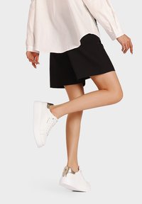 Sneakers bianchi con una superficie liscia, un accento dorato testurizzato sul tallone e una suola bianca spessa, abbinati a una gonna nera e una camicia bianca.