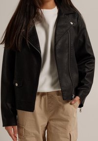 Donna che indossa una giacca da motociclista in pelle nera sopra una camicia bianca e pantaloni cargo beige, con una mano in tasca, su uno sfondo neutro.