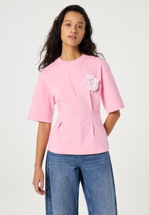 Woman with long dark hair wearing a pink shirt with a fabric flower detail and blue jeans, standing against a plain light background.