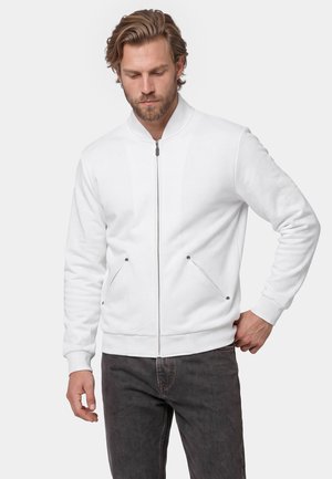 Man with beard wearing white zip-up jacket and dark jeans, standing with one hand on hip against plain background.