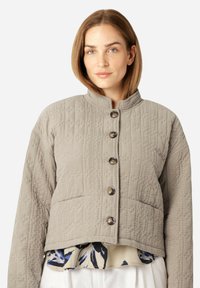 Grau gesteppte Jacke mit hohem Kragen, ausgestattet mit fünf metallischen Knöpfen, strukturierter Oberfläche und einem cropped Design, über einer blumigen Bluse getragen.