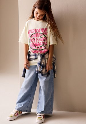 Noor tüdruk kannab üle suurust Nirvana T-särki, laiu helesiniseid teksapükse, ruudulist särk vöökohas seotud ja värvilisi tossusid, seisab pea alla kaldu.