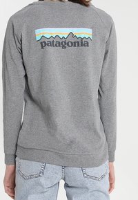 Grauer Pullover mit Rundhalsausschnitt, ausgestattet mit einem bedruckten Logo und einer Berggrafik in Blau, Orange und Türkis auf der Rückseite. Weiches Material.