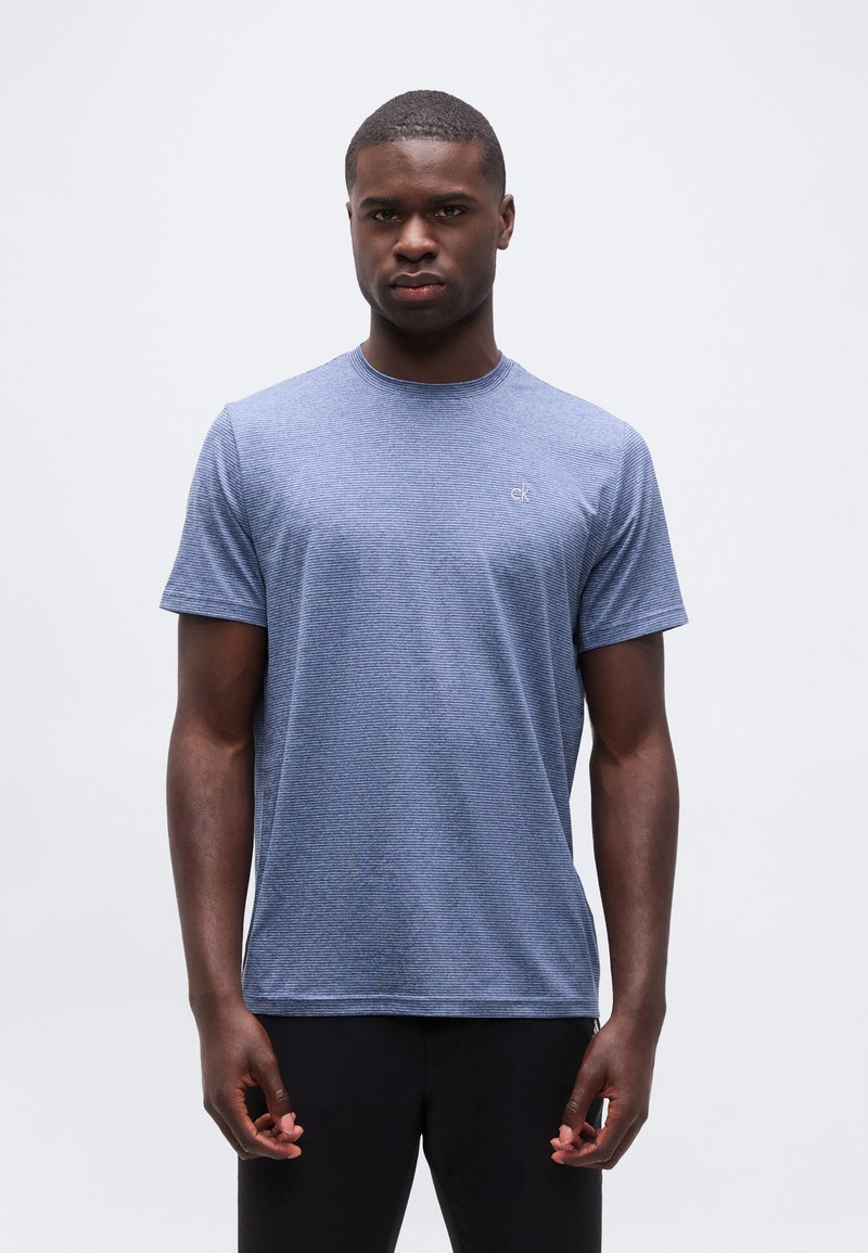 Άνδρας που στέκεται στραμμένος προς τα εμπρός φορώντας μπλε ριγέ κοντομάνικη μπλούζα και μαύρο παντελόνι, με φόντο ένα απλό φωτεινό φόντο.