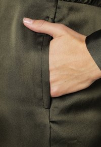 Tessuto verde oliva con una superficie liscia e satinata; posizionato a mano in una tasca laterale con un bordo pulito e una leggera lucentezza.