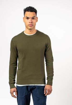 Olivgrüner Strickpullover mit Rundhalsausschnitt, langen Ärmeln und gerippten Bündchen. Wird über einem weißen Hemd getragen, kombiniert mit blauen Hosen.