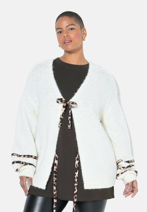 Weißer Strickcardigan mit weiten Ärmeln, verziert mit Tiermuster-Bindebändern an Taille und Bündchen, getragen über einem braunen Hemd. Weiche Textur.