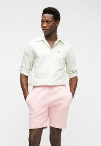 Άνδρας που στέκεται με τα χέρια στις τσέπες, φοράει ανοιχτοπράσινο πουκάμισο με κουμπιά και ροζ σορτς, σε απλό λευκό φόντο.