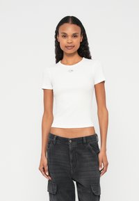 Fehér, ujjatlan póló bordázott textúrával és kerek nyakkivágással, amelyen egy kis ezüst embléma látható a mellkasnál, sötét cargo nadrággal párosítva.