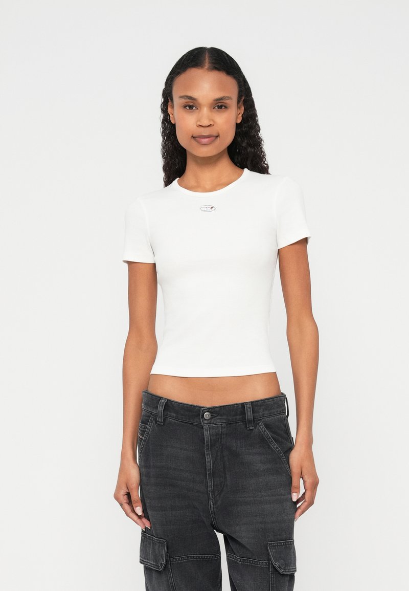 Fehér, ujjatlan póló bordázott textúrával és kerek nyakkivágással, amelyen egy kis ezüst embléma látható a mellkasnál, söt�ét cargo nadrággal párosítva.