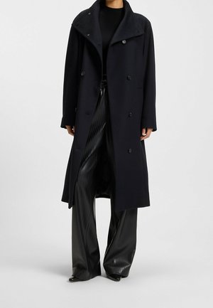 Persoon draagt een lange zwarte dubbele rij jassen over een zwarte top en zwarte wijde geplooide broek tegen een effen achtergrond.