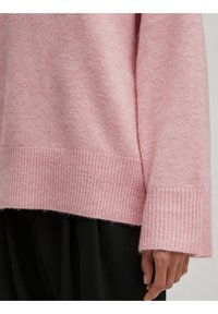 Hellrosa gestrickter Pullover mit geripptem Saum und Bündchen, bietet eine weiche Textur und eine lockere Passform. Über einem schwarzen Kleidungsstück getragen.