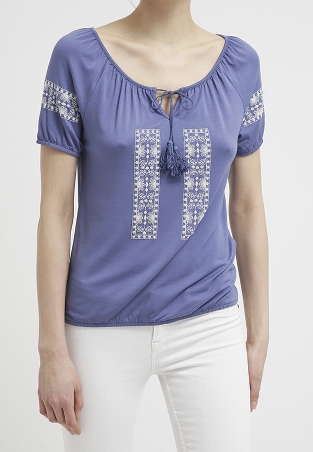 Haut court violet à manches courtes avec un décolleté en scoop, orné de broderies blanches décoratives et de petits pompons à l'avant, associé à un pantalon blanc.