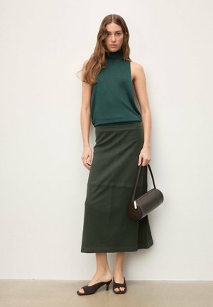 Haut vert sans manches et jupe midi vert foncé, avec une texture lisse. Sac à main rond marron avec une bandoulière et chaussures à talons ouverts.