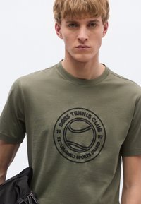 Olíva zöld pamut póló fekete kör alakú teniszlogóval és szöveggel. Rövid ujjú, kerek nyakú kialakítás.