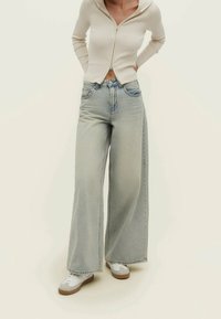 Light blue wide-leg jeans with a high waist and distressed detailing, paired with a cream ribbed zip-up hoodie. White sneakers complete the look.