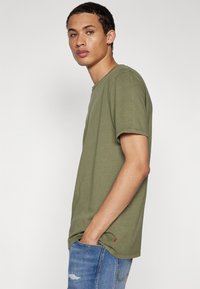 Oliwkowa koszulka z okrągłym dekoltem wykonana z bawełny. Krótkie rękawy, luźny krój, a po boku ma małą czerwoną metkę z logo. W zestawie z niebieskimi dżinsami.