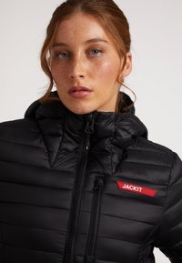 Giacca a vento nera con design a righe orizzontali, zip frontale, colletto alto e logo rosso "JACKIT" sul petto. Texture liscia.