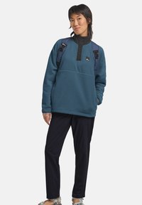 Pullover in pile color turchese con colletto nero a bottoni, indossato con pantaloni blu navy e uno zaino. Il design presenta una toppa con logo sul petto.