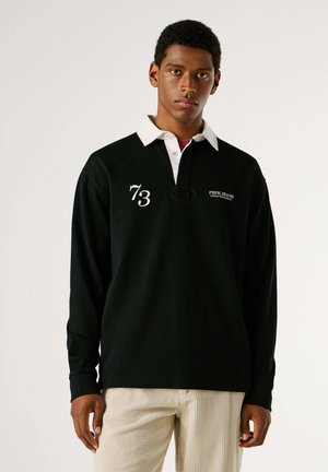 Camiseta polo negra de manga larga con cuello blanco. Presenta un logo y el número "73". Combinada con pantalones de pana de color claro. Textura suave.