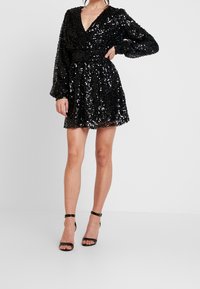 Kvinna klädd i en svart långärmad miniklänning täckt av paljetter och svarta högklackade sandaler med ankelsnodd, stående på ett vitt golv.