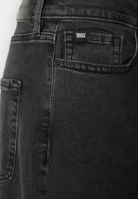 Juodi džinsai su klasikinio sukirpimo, minimalistiniu dizainu ir mažu DKNY džinsų etikete ant nugarinės kišenės. Tekstūruota medžiaga su vos pastebimu blizgesiu.