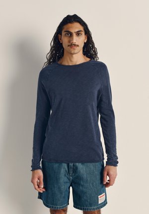Camisa de manga larga azul marino con cuello redondo y mangas raglán, hecha de una tela suave. Combinada con pantalones cortos de mezclilla con un parche de logo.