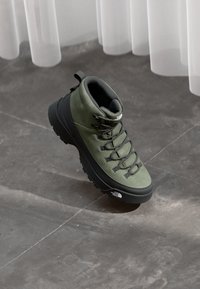 Green hiking boot with suede upper, black laces, rugged rubber sole, and a notable logo on the side. Curved design and padded collar.
