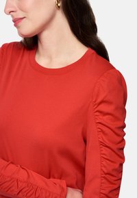 Red long-sleeve top with a round neckline, featuring gathered fabric details on the sleeves and a smooth, soft texture.