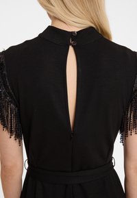 Zwarte jurk met een hoge kraag, opening aan de achterkant met sleutelgat en knoopsluiting. Heeft gemeleerde, franjesmouwen met kralenaccenten.