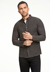 Camicia button-up grigio scuro realizzata in tessuto testurizzato, con tasca sul petto e orlo tondo, indossata sopra una maglietta bianca. Pantaloni neri.