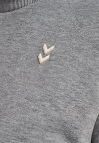 Jasnoszara tkanina z dwoma małymi kremowymi wyhaftowanymi szewronami blisko górnego środka.