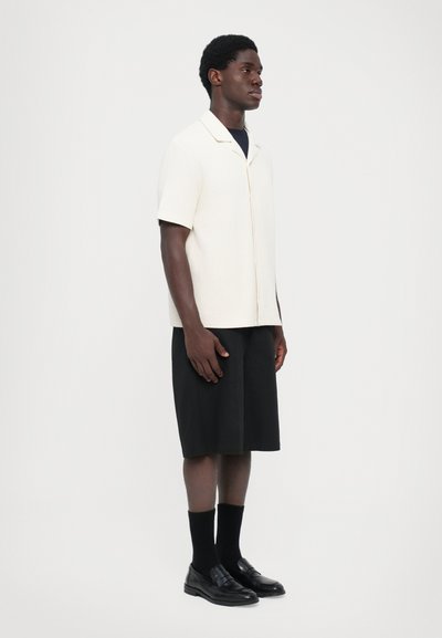 Cremefarbenes Kurzarmhemd mit lockerer Passform, kombiniert mit schwarzen knielangen Shorts und schwarzen Slippern, die ein minimalistisches Design präsentieren.