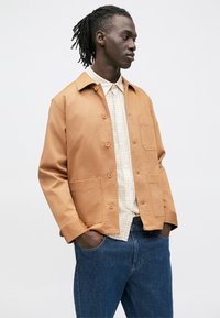 Brun bomullsjakke med to frontlommer, knappestenging og avslappet passform, båret over en lys rutete skjorte og blå jeans.