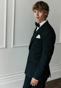 Schwarzer Smoking mit satinierter Revers, ausgestattet mit einem weißen Taschentuch und klassischer Fliege. Stilvoll mit taillierter Passform und strukturierten Details.