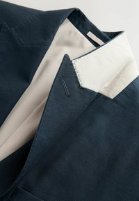 Tmavomodré sako so svetlobéžovým vnútorným podšívaním, zobrazujúce prešívaný klopový golier a detail goliera.