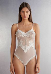 Spitzenbodysuit in Elfenbein mit einem gewellten Ausschnitt und floralem Muster, ausgestattet mit dünnen Trägern und einem transparenten Netzgewebe.
