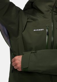 Giubbotto impermeabile verde con cappuccio, caratterizzato dal logo Mammut, diverse tasche con zip e polsini regolabili. Tessuto liscio e durevole.