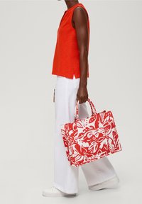 Červená a bílá taška přes rameno se silnými uchy, obdélníkový tvar, látkový materiál a květinový vzor. Nesená s červeným topem a bílými kalhotami.