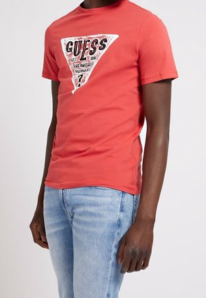 Homme portant un t-shirt rouge corail avec un logo triangulaire blanc et le texte noir "GUESS Los Angeles California", assorti à un jean bleu clair.