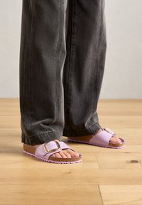 Persona che indossa pantaloni larghi grigi scuri e ciabatte lavanda con grandi fibbie dorate, in piedi su un pavimento di legno.