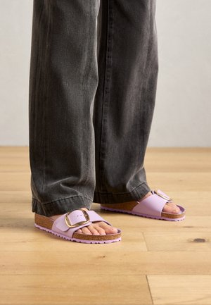 Person wearing loose dark gray pants and lavender slide sandals with large gold buckles standing on wooden floor.