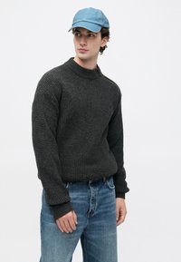 Junger Mann, der einen strukturierten dunkelgrauen Pullover, hellblaue Jeans und eine hellblaue Baseballmütze trägt, und seitlich vor einem schlichten Hintergrund schaut.