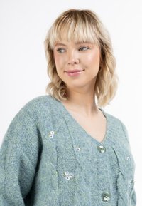 Texturerad grön cardigan med V-halsringning, prydd med broderade vita blommor och stora gröna knappar fram. Mjuk, bekväm stickad material.