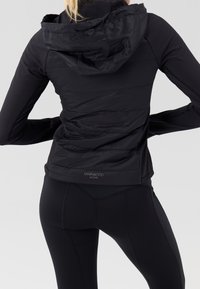 Chaqueta deportiva negra con capucha, con textura acolchada en la parte superior de la espalda y mangas largas ajustadas, combinada con leggings negros de talle alto.